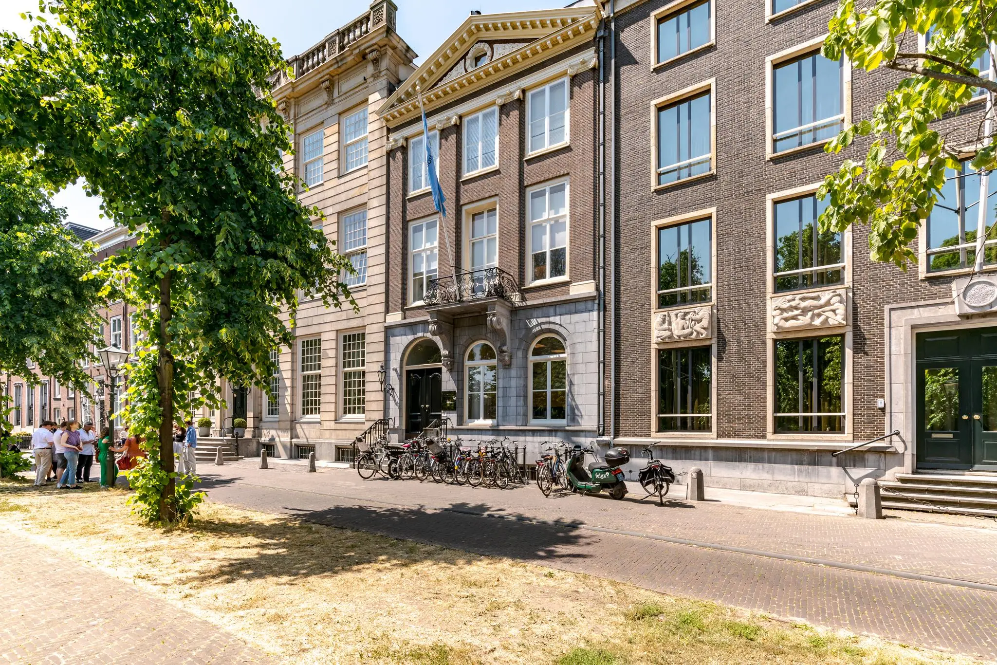 Zonnige straat aan de Lange Vijverberg in Den Haag met historische gebouwen, fietsen en een groepje mensen onder de bomen.